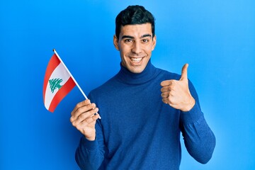 Handsome hispanic man holding lebanon flag smiling happy and positive, thumb up doing excellent and approval sign