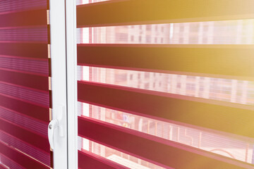 Evening sunlight enters the room through the blinds. Details of red fabric roller blinds on the plastic window in the room or kitchen. Window roller, duo system day and night. Dual roller shades  © Oleksandr