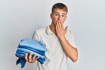 Young caucasian man holding stack of folded jeans covering mouth with hand, shocked and afraid for...