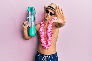 Young caucasian man wearing swimsuit and hawaiian lei holding watergun with open hand doing stop...