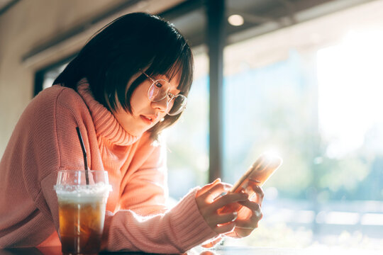 Young adult asian woman using mobile phone for social media background with window and warm sunlight on winter