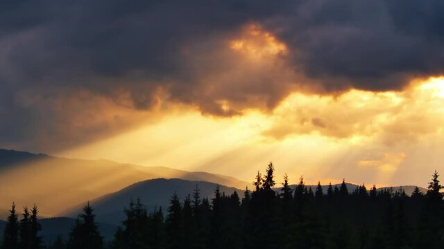 Time-lapse of sun rays emerging though the dark storm clouds in the mountains with a forest on a foreground
