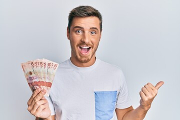 Handsome caucasian man holding 10 colombian pesos banknotes pointing thumb up to the side smiling...