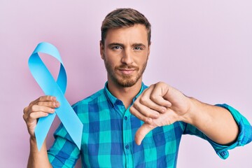 Handsome caucasian man holding blue ribbon with angry face, negative sign showing dislike with thumbs down, rejection concept