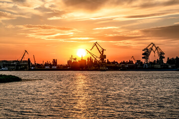 Kherson port at sunset (Ukraine). View from the Dnieper River to the coastline with harbor cranes and commercial ships illuminated by orange sunbeams