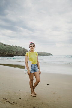 plano entero de mujer joven posando con pantalones cortos remera amarilla y gafas de sol,  posando en la playa mirando hacia la camara