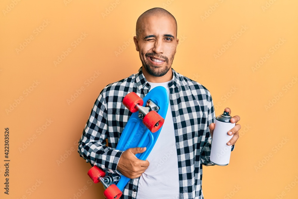 Poster Hispanic adult skater man holding skate and graffiti spray winking looking at the camera with sexy expression, cheerful and happy face.