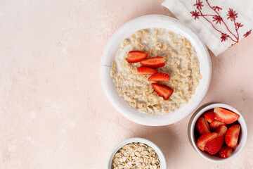 Oatmeal porridge with strawberries in a white plate on a linen napkin on pink background. Breakfast health food concept. Top view