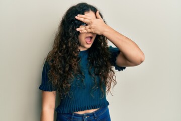 Young brunette woman with curly hair wearing casual clothes peeking in shock covering face and eyes with hand, looking through fingers with embarrassed expression.