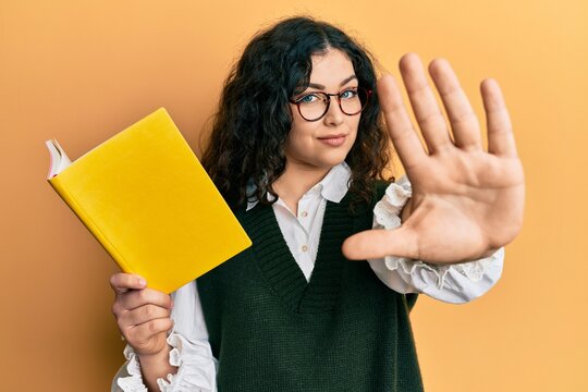 Young brunette woman with curly hair reading a book wearing glasses with open hand doing stop sign with serious and confident expression, defense gesture