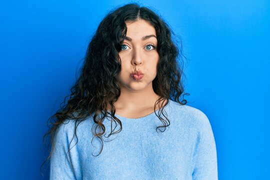Young brunette woman with curly hair wearing casual clothes puffing cheeks with funny face. mouth inflated with air, crazy expression.