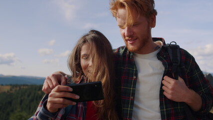 Smiling guy and girl looking video on smartphone screen during hike in mountains