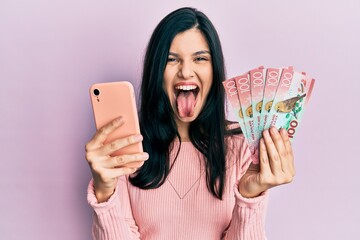 Young hispanic woman using smartphone holding new zealand dollars banknotes sticking tongue out happy with funny expression. © Krakenimages.com