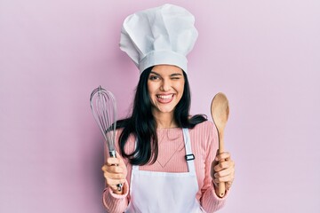 Young hispanic woman wearing baker uniform holding spoon and whisk winking looking at the camera...