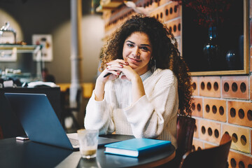 Portrait of skilled female student with education textbook sitting at cafeteria table with modern netbook technology and looking at camera, millennial copywriter with digital laptop computer posing