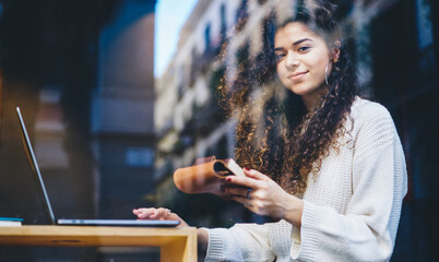 Portrait of skilled female student with education textbook in hand sitting at cafeteria table with modern netbook technology and looking at camera, window view on millennial copywriter with notepad