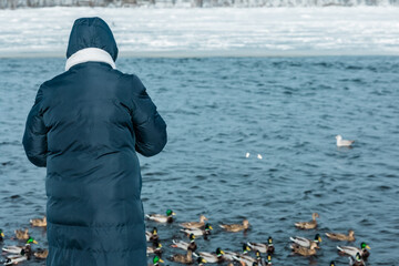 A woman in a blue jacket feeds ducks on the bank of a frozen river in snow and ice, on a sunny winter day.