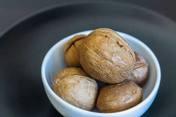 Heap, pile of whole walnuts in a round-shaped brown shell lie in small white platter on black plate on blurred bokeh background