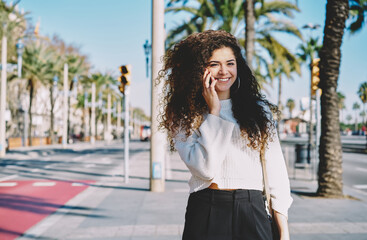 Portrait of cheerful female tourist with curly hair smiling at camera while using cellphone technology for friendly calling, happy girl enjoying international conversation for talking with family