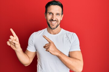 Young handsome man wearing casual white tshirt smiling and looking at the camera pointing with two hands and fingers to the side.