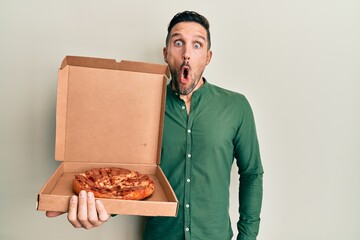 Handsome man with beard holding italian pizza scared and amazed with open mouth for surprise, disbelief face