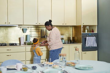 Back view at happy African-American mother and daughter cooking together in kitchen interior, copy space