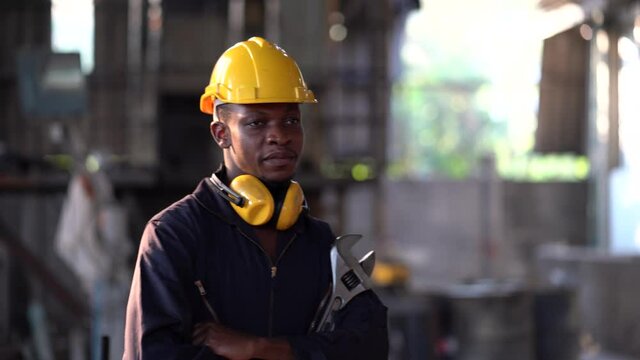 portrait of african american heavy industry worker wearing hard hat and yellow ear muffs standing confident arms crossed and in holding  wrench tools in Large Industrial Factory. young  black engineer