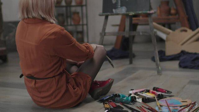 Inspired talented lovely female artist in creative process sitting cross legged on floor, looking at painting thoughtfully, surrounded by artistic tools, paints, paintbrushes and palettes in workshop.