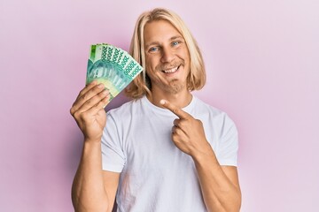 Caucasian young man with long hair holding 20000 indonesian rupiah smiling happy pointing with hand and finger