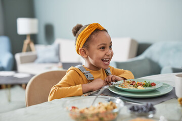 Portrait of cute African-American girl sitting at dinner table and laughing while enjoying meal in...
