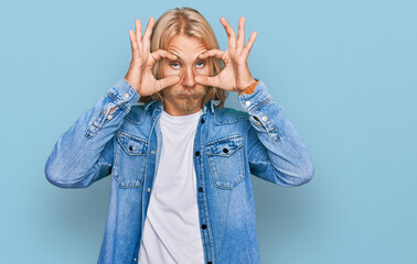 Caucasian man with blond long hair wearing casual denim jacket trying to open eyes with fingers,...
