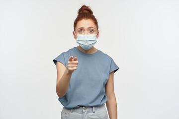 Portrait of shocked girl with ginger hair bun. Wearing blue t-shirt and medical, protective face mask. Pointing at you. Can't believe her eyes. Watching at the camera, isolated over white background