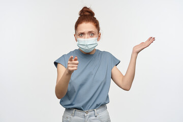 Confused, unsure woman with ginger hair bun. Wearing blue t-shirt and medical, protective face mask. Shrug and pointing at you, asking. Watching at the camera, isolated over white background