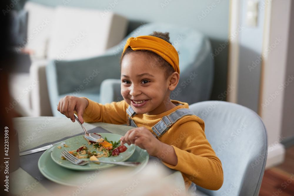 Wall mural portrait of cute african-american girl eating food at table and smiling happily while enjoying dinne