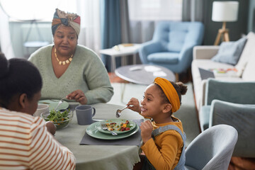Side view portrait of cute African-American girl eating food at table and smiling happily while enjoying dinner with family in cozy home interior