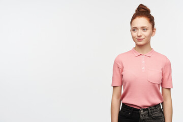 Young lady, pretty woman with ginger hair bun. Wearing pink t-shirt and black jeans. Confident smiling. Introducing herself. Watching to the left at copy space, isolated over white background