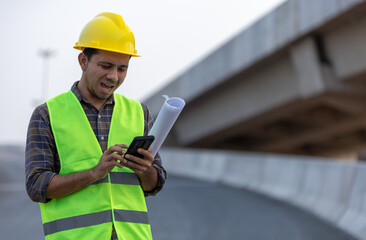 Asian construction workers use smartphone and hold a plan blueprint at the construction site.