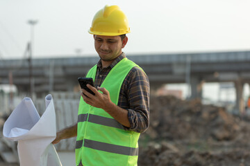 Asian construction workers use smartphone and hold a plan blueprint at the construction site.