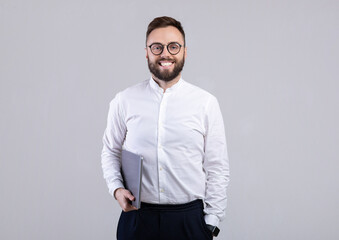 Happy young businessman in formal clothes and glasses holding laptop computer on grey studio background