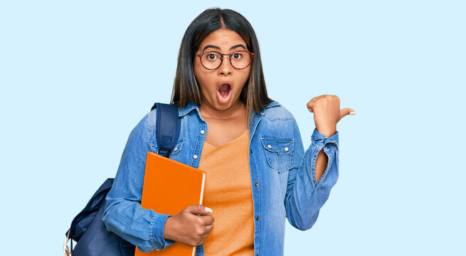 Young latin girl wearing student backpack and holding books surprised pointing with hand finger to the side, open mouth amazed expression.