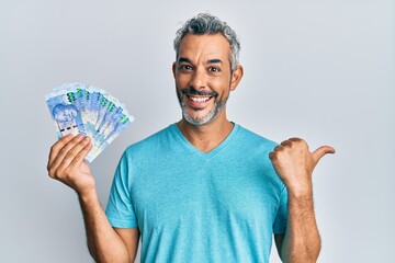 Middle age grey-haired man holding south african rands banknotes pointing thumb up to the side smiling happy with open mouth