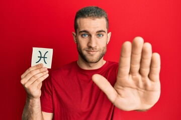 Young caucasian man holding paper with pisces zodiac sign with open hand doing stop sign with serious and confident expression, defense gesture