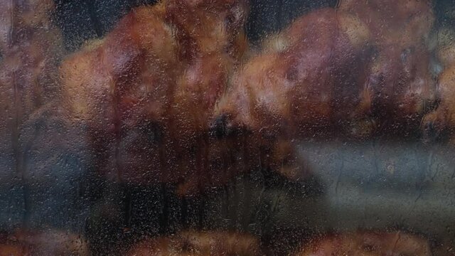 Close-up shot of a steamy rotisserie machine window with water drops. Grilled roasted whole chickens with tasty golden-yellow roasted skin on a turning spit in a row. Restaurant, butcher shop roaster.