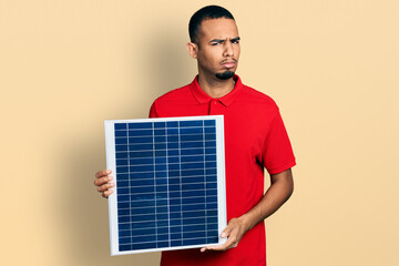 Young african american man holding photovoltaic solar panel skeptic and nervous, frowning upset...