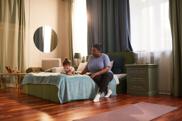 Candid full length portrait of African-American woman sitting on bed with daughter in cozy home...