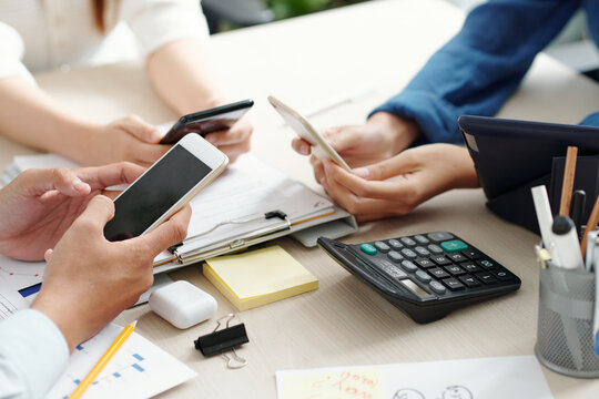 Hands of business people sitting at meeting table with charts and reports and using applications on smartphones