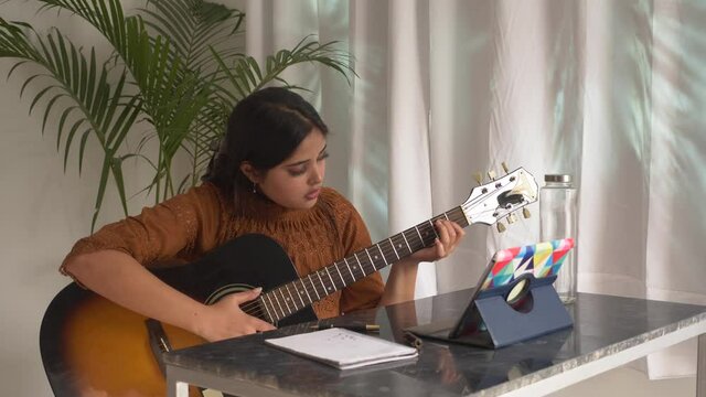 Young Asian woman practicing and learning how to play guitar online on laptop tablet.Female guitarist watching online guitar tutorial.Concept of online guitar classes, practicing at home.