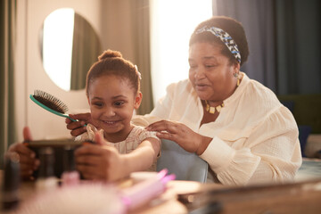 Portrait of happy African-American grandmother styling hair of cute little girl in cozy home setting