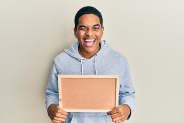 Young handsome hispanic man holding empty corkboard winking looking at the camera with sexy...