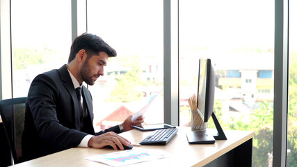 Happy young professional businessman sitting in office and working and looking at computer monitors and documents in the workplace. The concept for data analytics and online technology